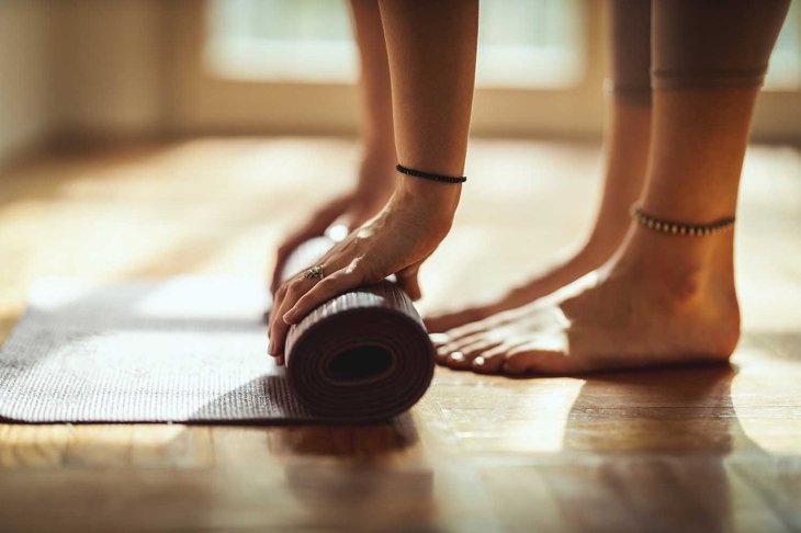 Close up of a womans hands is rolling up exercise mat and preparing to doing yoga. She is exercising on floor mat in morning sunshine at home.