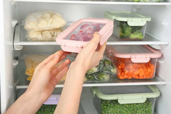 Woman putting container with tomatoes in refrigerator with frozen vegetables, closeup